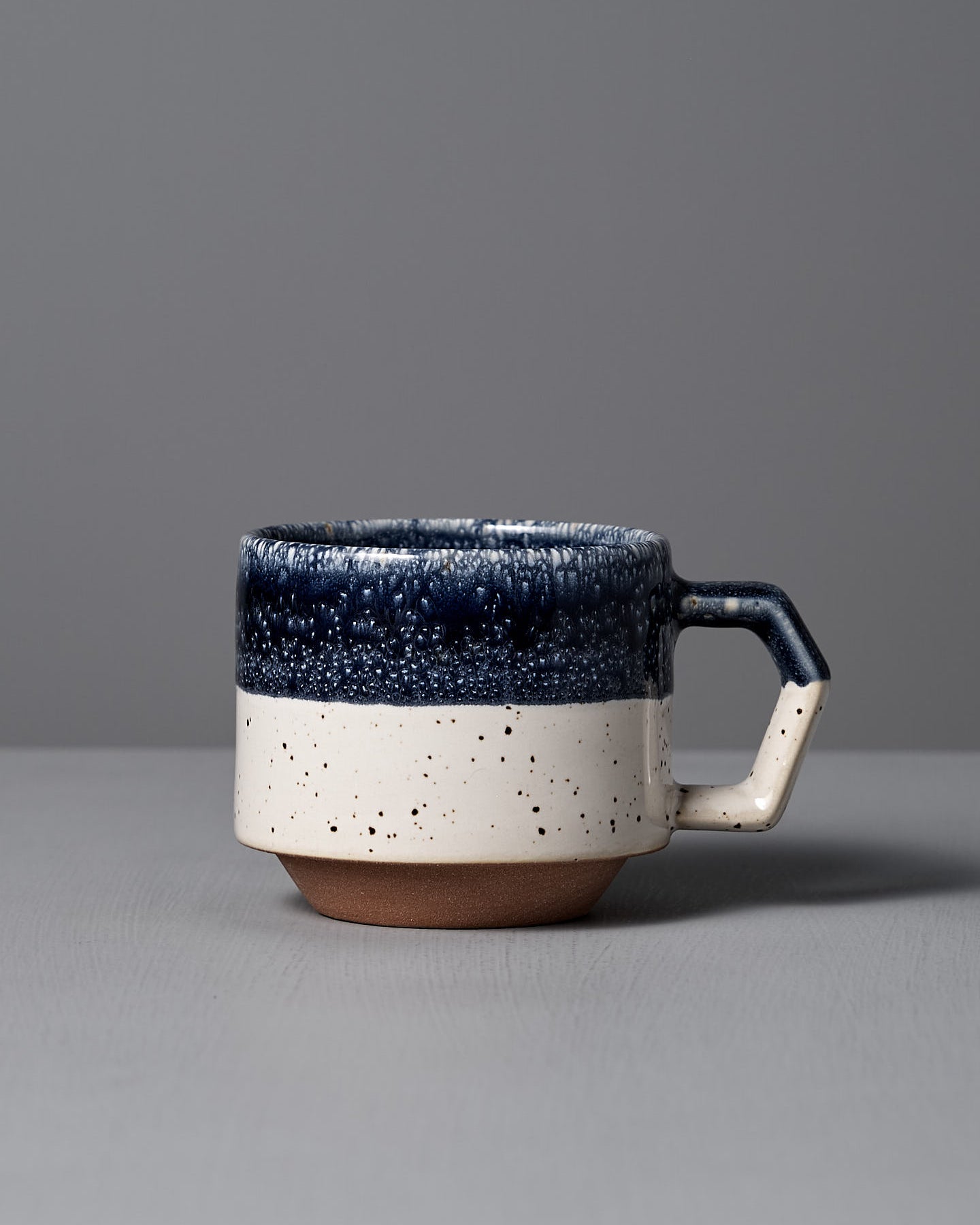 The Stacking Mug – White-Navy Drop by CHIPS Inc. features elegant Minoyaki porcelain with a textured dark blue top, speckled white bottom, and square handle. Its exquisite craftsmanship and stackable design make it a perfect collection addition, beautifully placed on any gray surface.
