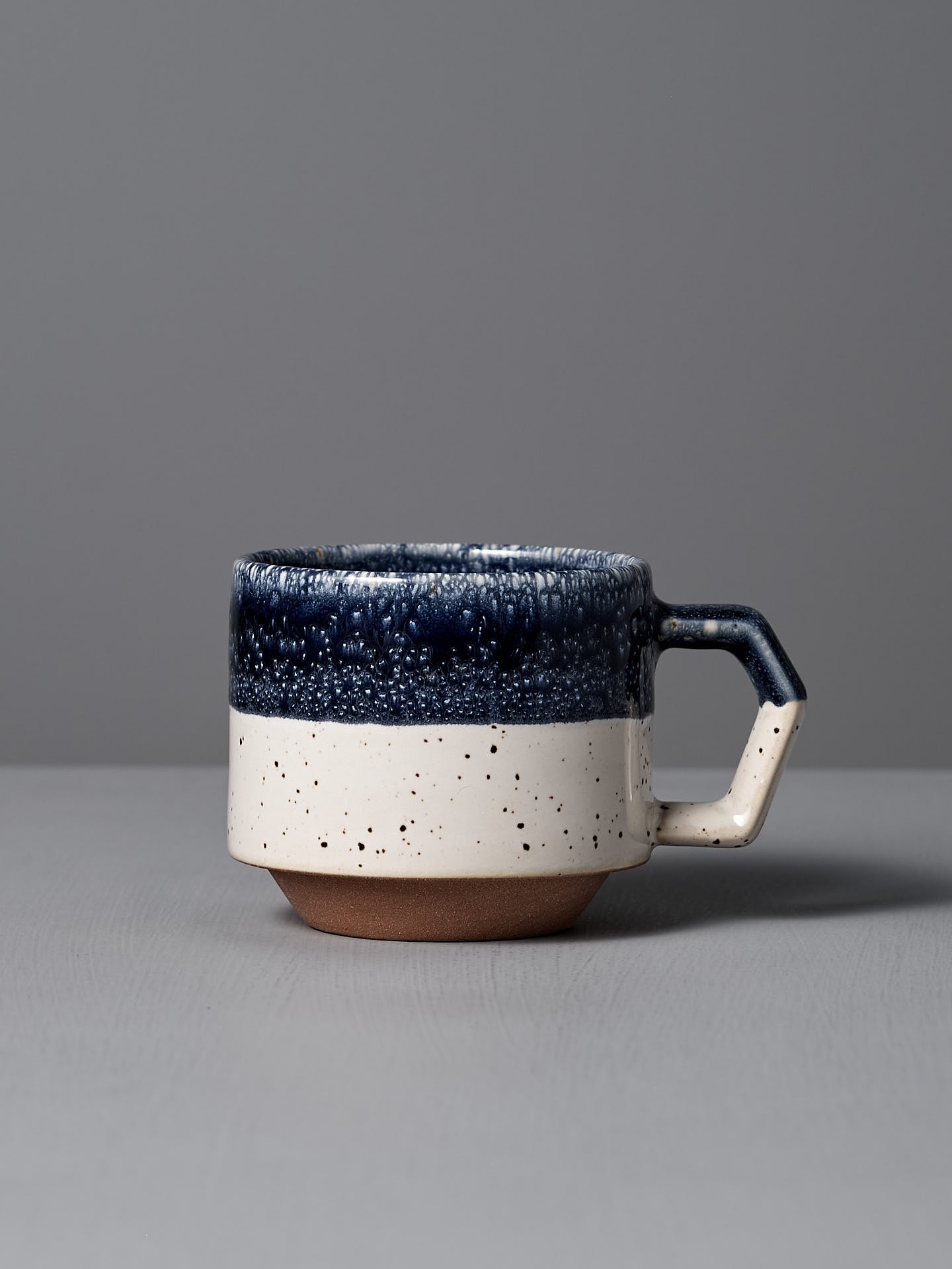 The Stacking Mug – White-Navy Drop by CHIPS Inc. features elegant Minoyaki porcelain with a textured dark blue top, speckled white bottom, and square handle. Its exquisite craftsmanship and stackable design make it a perfect collection addition, beautifully placed on any gray surface.