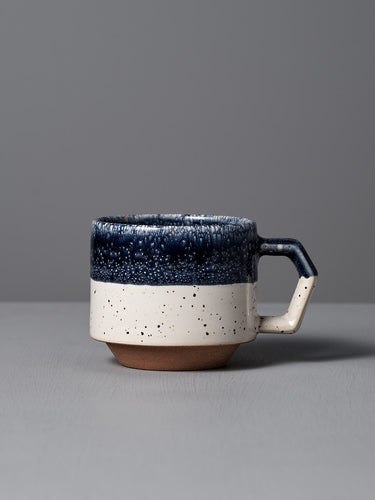 The Stacking Mug – White-Navy Drop by CHIPS Inc. features elegant Minoyaki porcelain with a textured dark blue top, speckled white bottom, and square handle. Its exquisite craftsmanship and stackable design make it a perfect collection addition, beautifully placed on any gray surface.