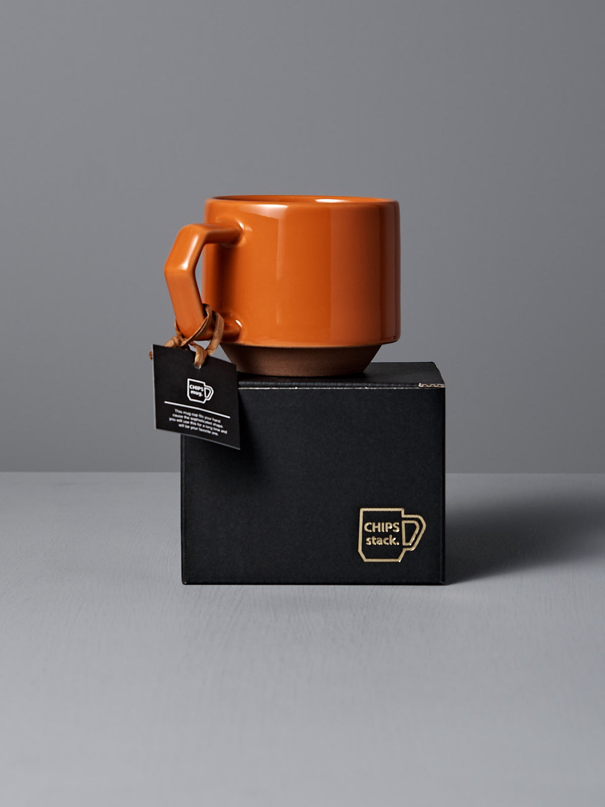 A stylish orange mug with a rectangular handle rests atop a black box labeled &quot;CHIPS stack.&quot; Made from Minoyaki porcelain, this handmade piece from Japan is enhanced by a tag hanging from the handle, exuding elegance.