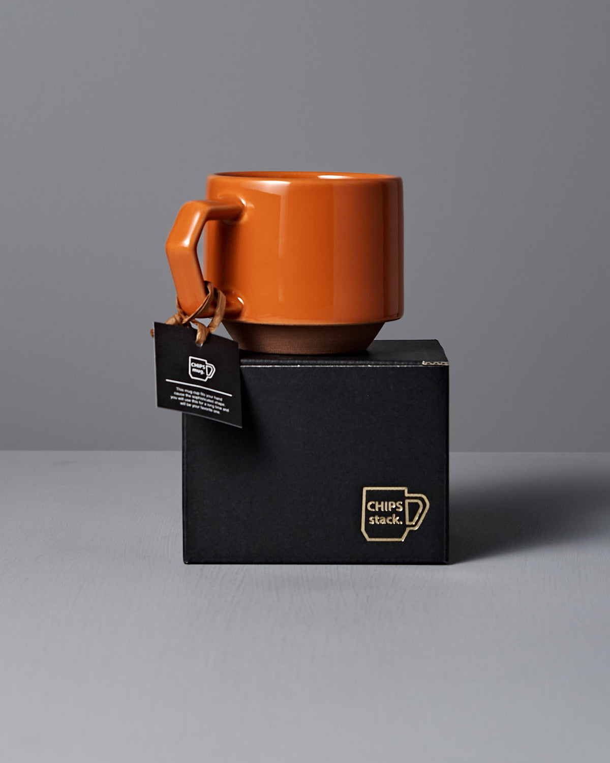 A stylish orange mug with a rectangular handle rests atop a black box labeled "CHIPS stack." Made from Minoyaki porcelain, this handmade piece from Japan is enhanced by a tag hanging from the handle, exuding elegance.