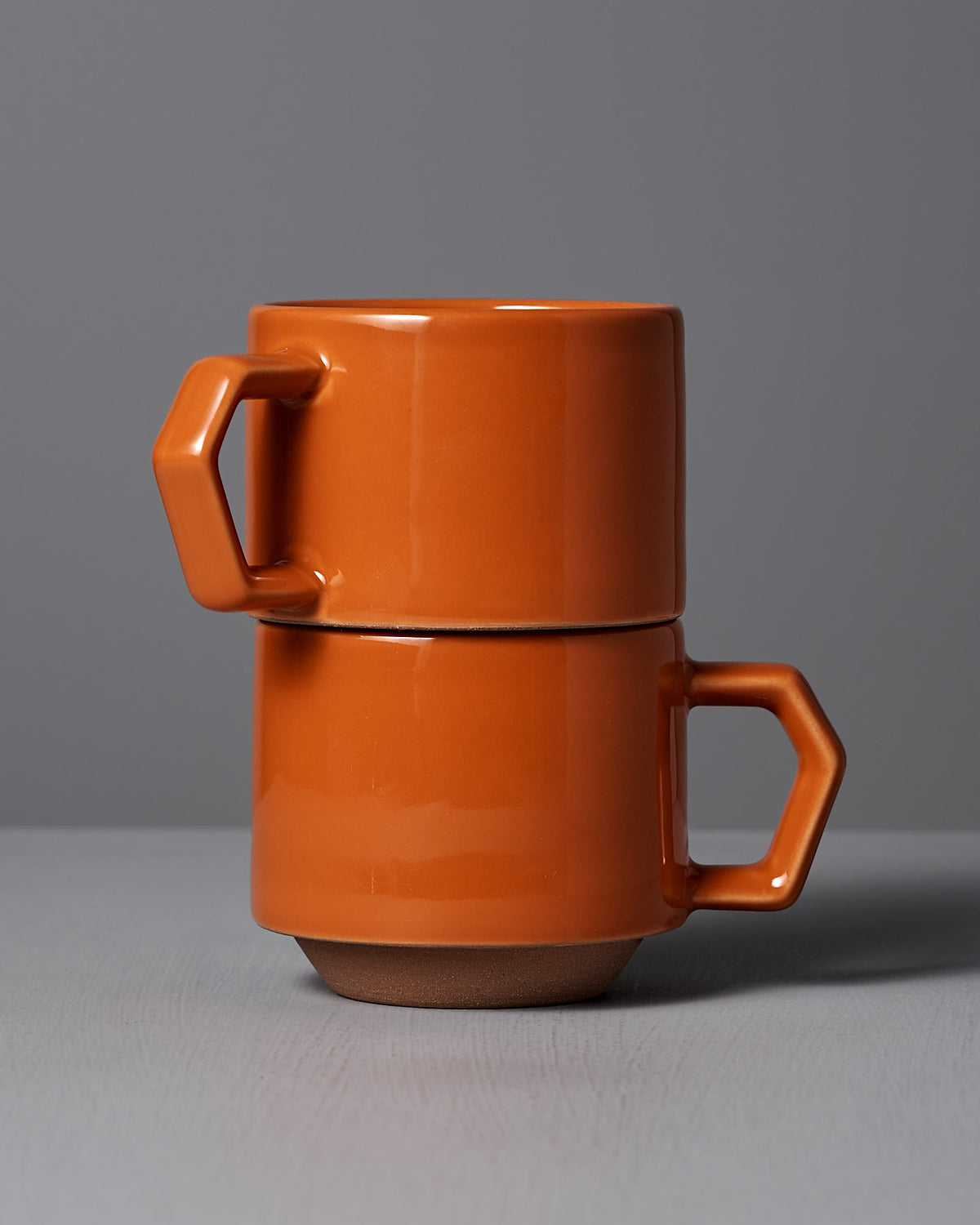 Two stylish stacking mugs from CHIPS Inc. in an orange Minoyaki porcelain are beautifully arranged on a gray surface against a matching background, highlighting their refined beauty.