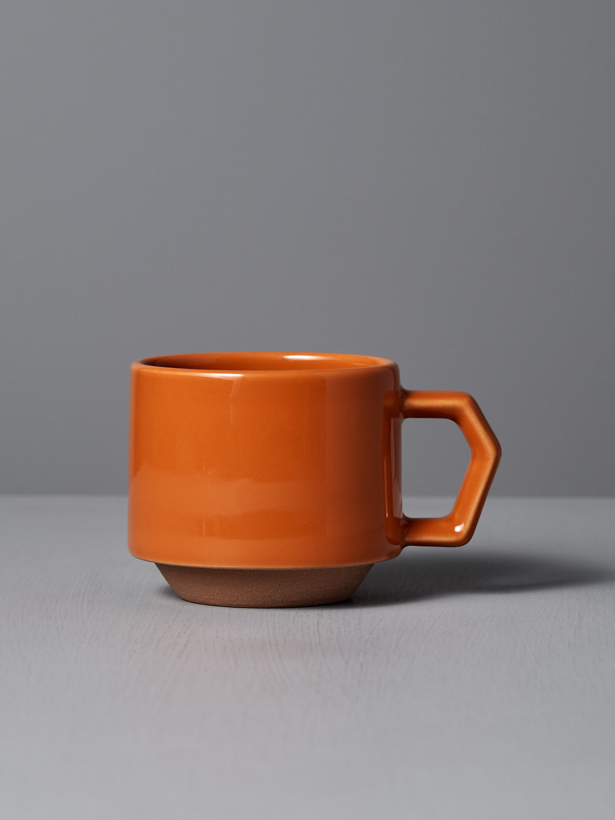 A sophisticated Stacking Mug – Orange made from Minoyaki porcelain, featuring a hexagonal handle and crafted by CHIPS Inc. artisans, is set against a gray surface and background.