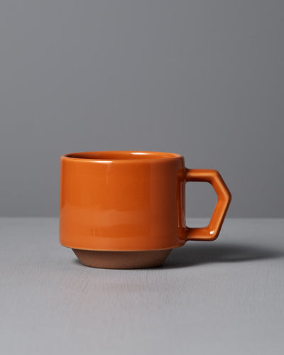 A sophisticated Stacking Mug – Orange made from Minoyaki porcelain, featuring a hexagonal handle and crafted by CHIPS Inc. artisans, is set against a gray surface and background.