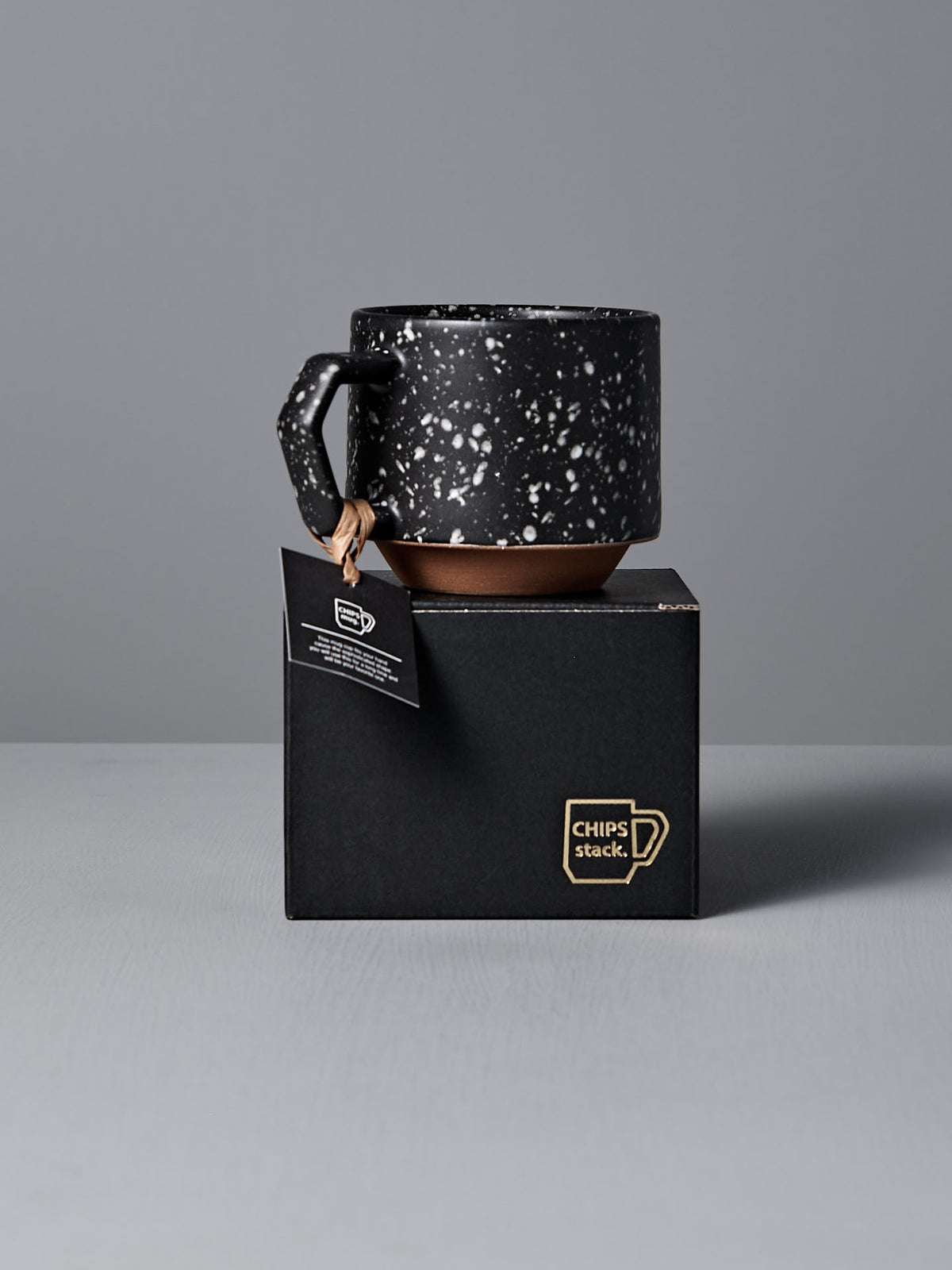 A Stacking Mug – Speckled Black by CHIPS Inc. rests on a black box with the label &quot;CHIPS stack,&quot; placed against a plain gray background.