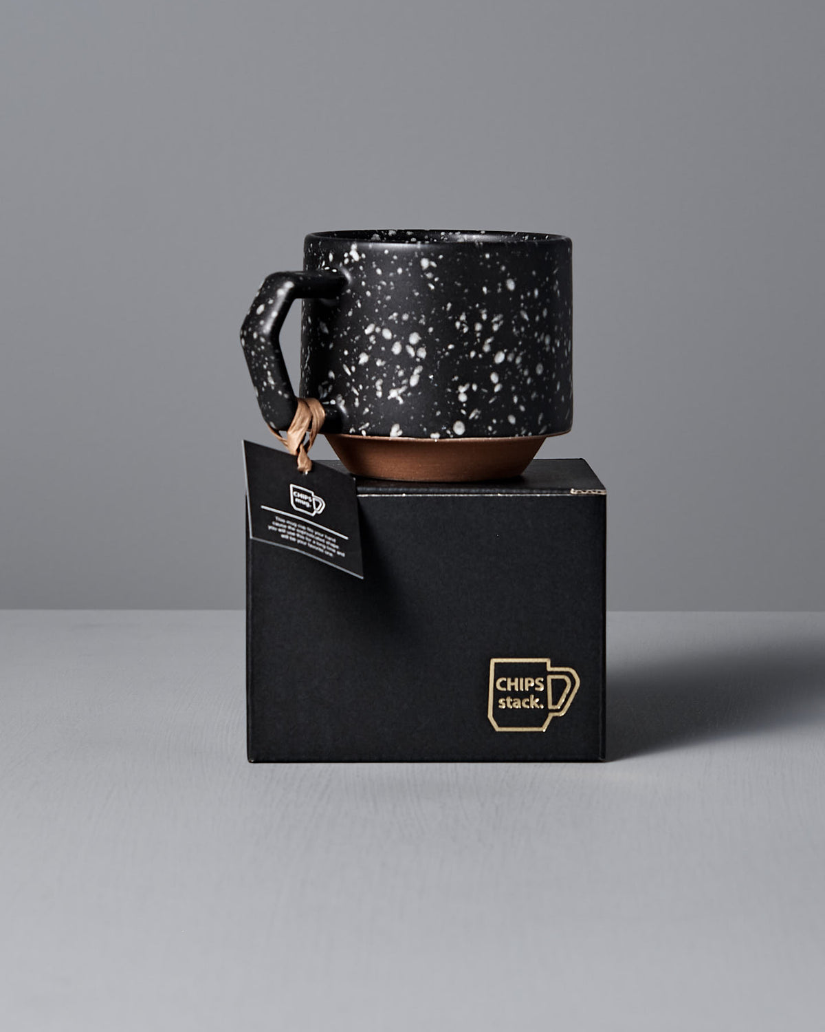 A Stacking Mug – Speckled Black by CHIPS Inc. rests on a black box with the label "CHIPS stack," placed against a plain gray background.