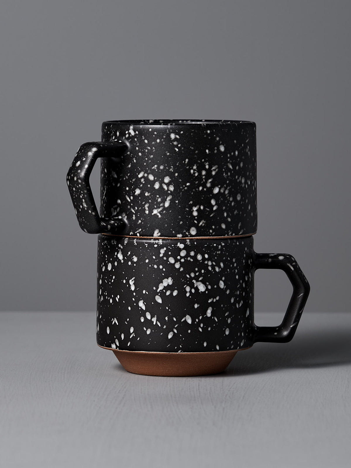 Two CHIPS Inc. Stacking Mugs in Speckled Black are elegantly stacked on a gray surface against a matching gray background, highlighting their sleek design and chip-free durability.