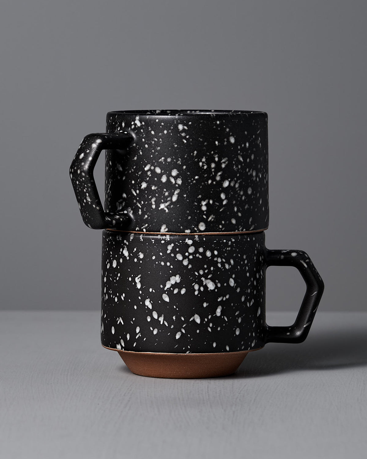 Two CHIPS Inc. Stacking Mugs in Speckled Black are elegantly stacked on a gray surface against a matching gray background, highlighting their sleek design and chip-free durability.