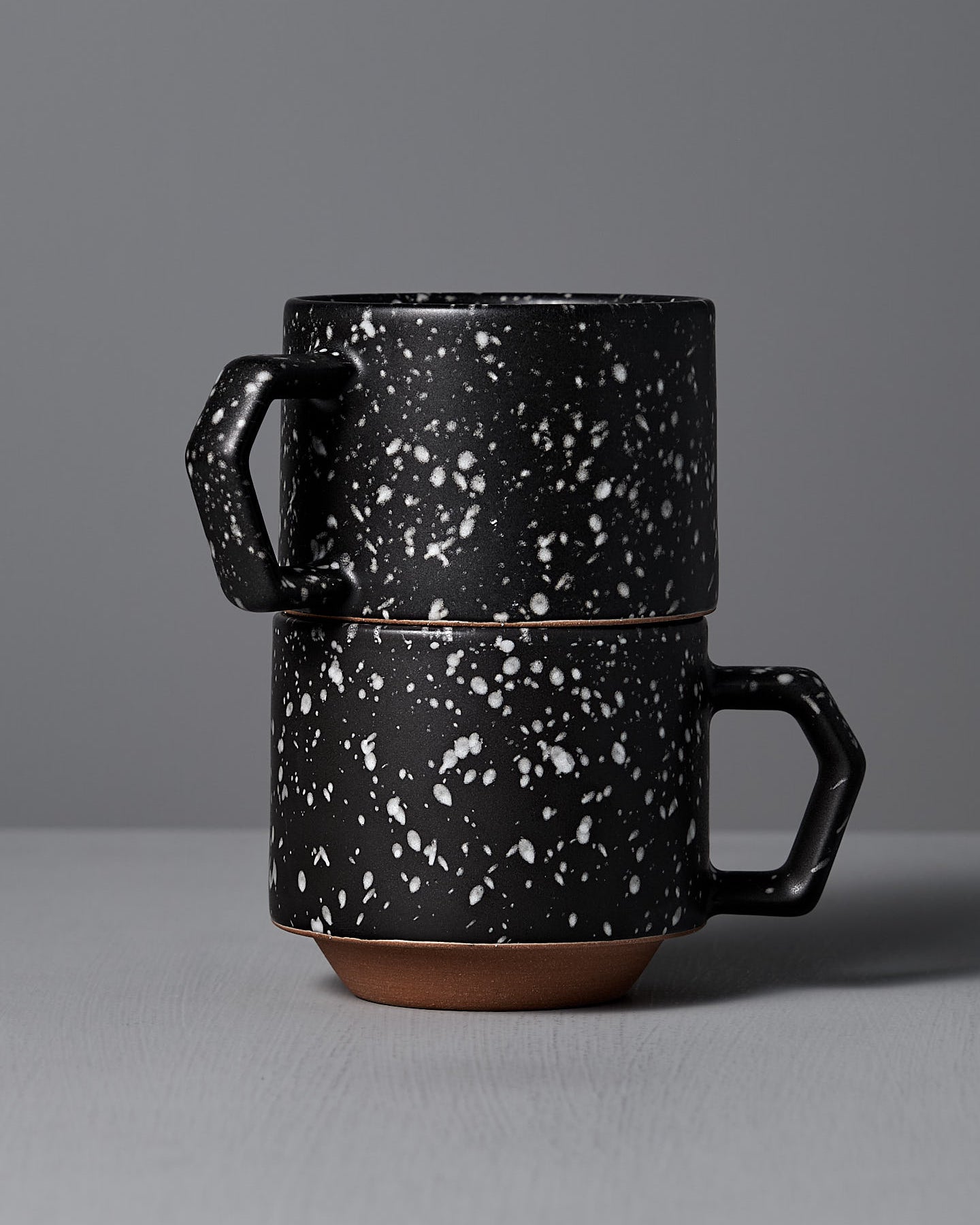 The Stacking Mug – Speckled Black by CHIPS Inc., elegantly crafted from Minoyaki porcelain, showcases a black ceramic body with white speckles and an angular handle, resting gracefully on a gray surface.