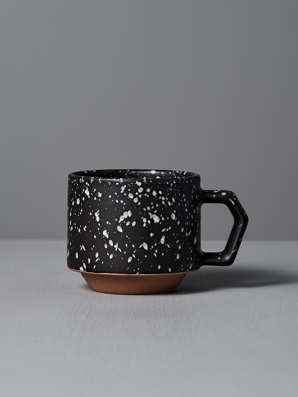 The Stacking Mug – Speckled Black by CHIPS Inc., elegantly crafted from Minoyaki porcelain, showcases a black ceramic body with white speckles and an angular handle, resting gracefully on a gray surface.