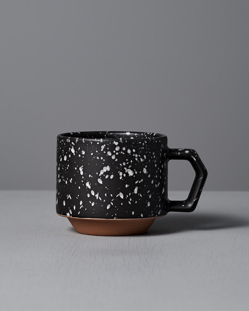 The Stacking Mug – Speckled Black by CHIPS Inc., elegantly crafted from Minoyaki porcelain, showcases a black ceramic body with white speckles and an angular handle, resting gracefully on a gray surface.