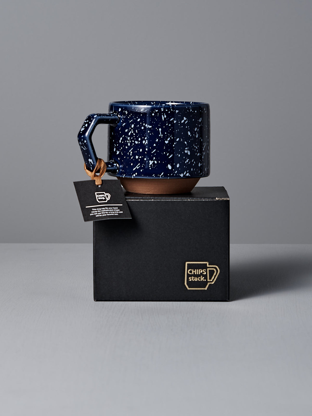 A Stacking Mug – Speckled Navy by CHIPS Inc., featuring a square handle, sits elegantly on a black box with a small tag attached to the handle.