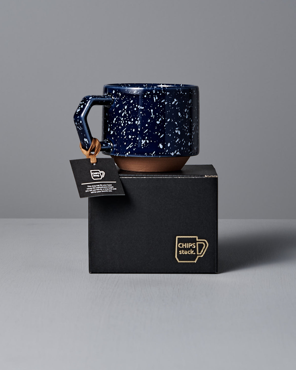 A Stacking Mug – Speckled Navy by CHIPS Inc., featuring a square handle, sits elegantly on a black box with a small tag attached to the handle.