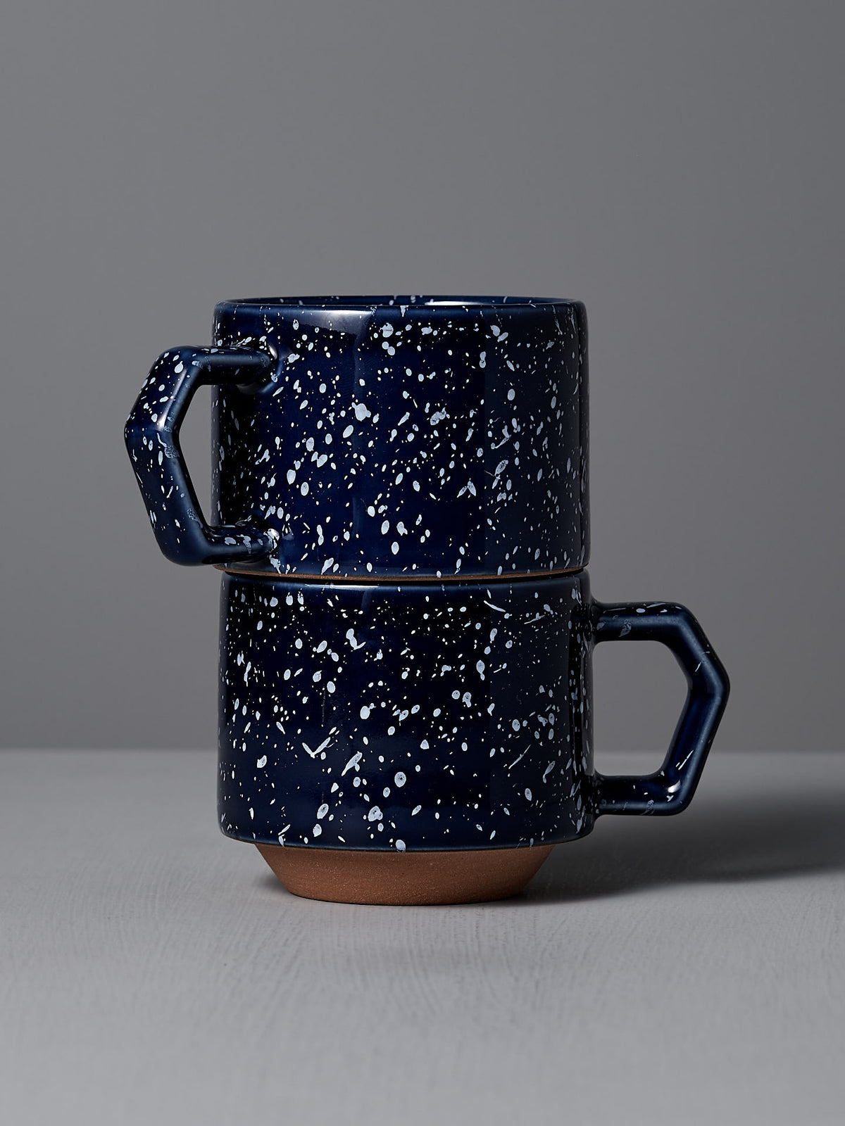 Two CHIPS Inc. Stacking Mugs in speckled navy, crafted from elegant Minoyaki porcelain, showcase a stackable design on a gray surface against a light gray background.