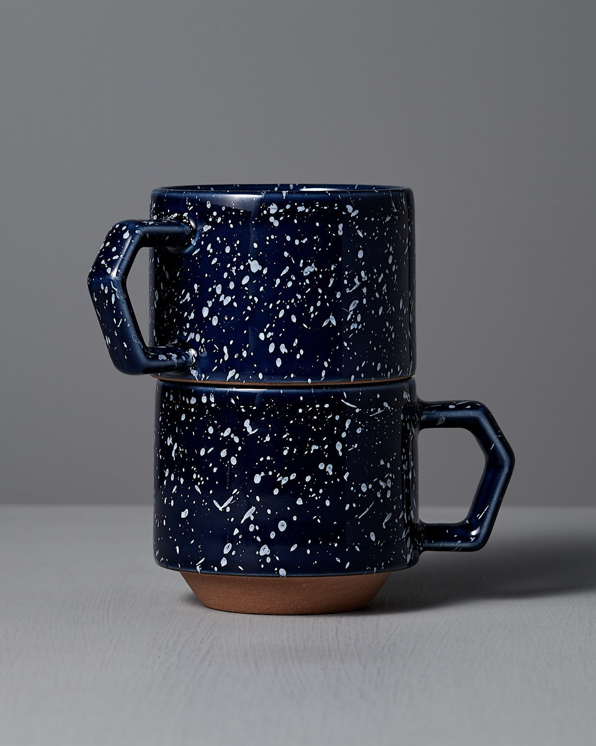 Two CHIPS Inc. Stacking Mugs in speckled navy, crafted from elegant Minoyaki porcelain, showcase a stackable design on a gray surface against a light gray background.
