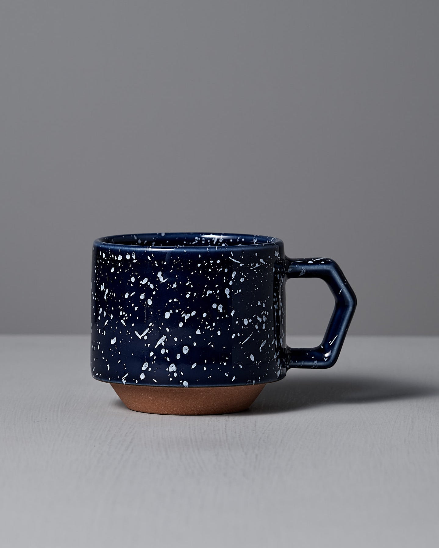 A Stacking Mug in speckled navy by CHIPS Inc. features a hexagonal handle and sits elegantly on a light gray surface, highlighting its stackable design.