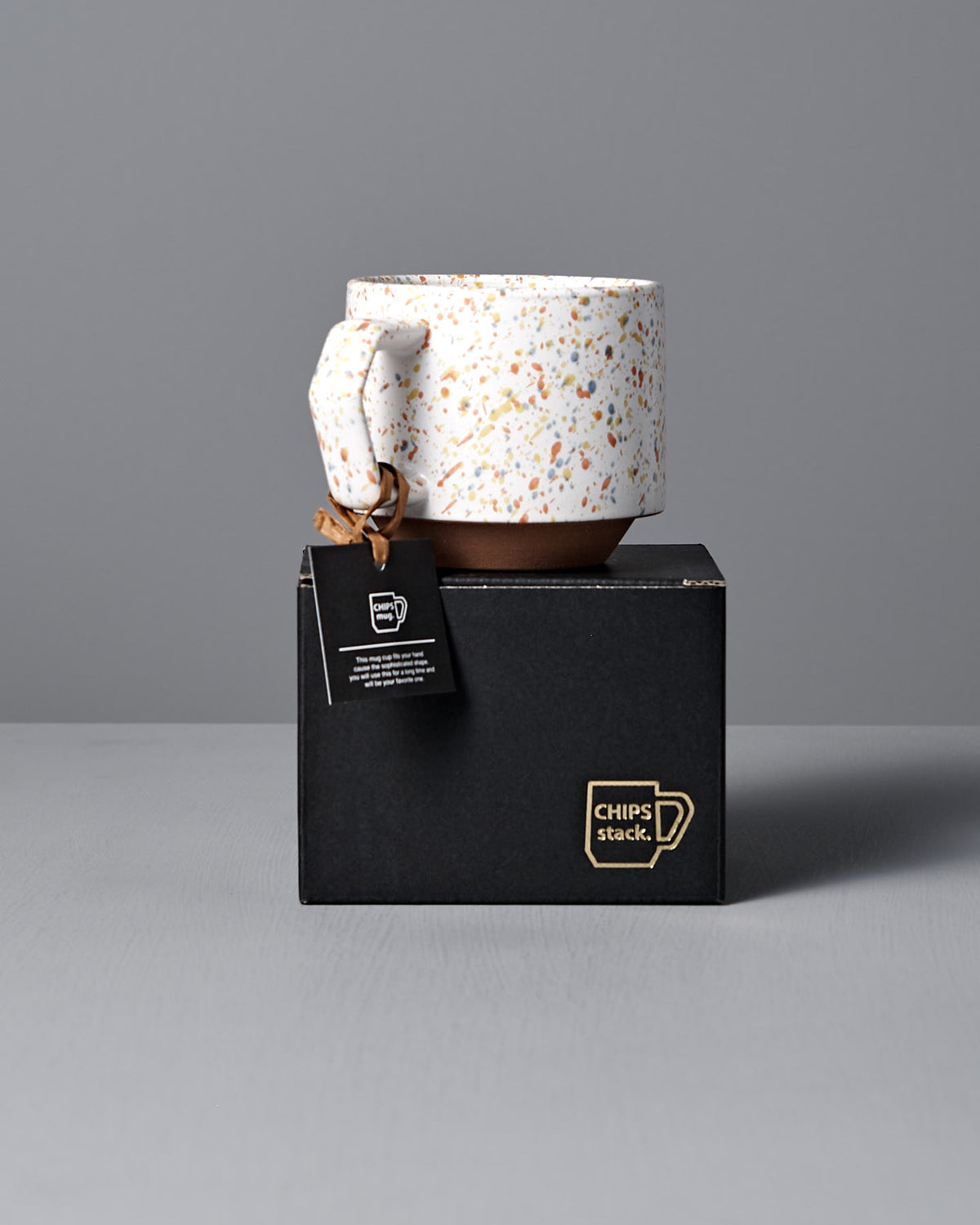 A speckled ceramic mug with a handle rests on a black box featuring the "CHIPS Inc." logo. Reminiscent of Japanese tableware, this elegant Stacking Mug – Speckled White exudes timeless charm. A small black tag attached to the handle hints at its sophisticated heritage.