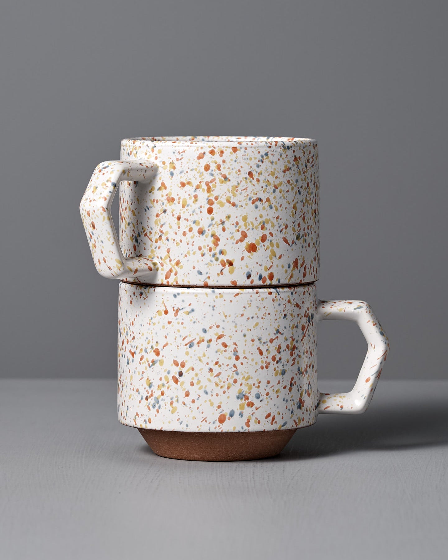 The Stacking Mug – Speckled White, a beautifully crafted piece by CHIPS Inc. artisans, boasts an elegant geometric handle. It is made from speckled Minoyaki porcelain and sits gracefully on a gray surface against a matching gray background.