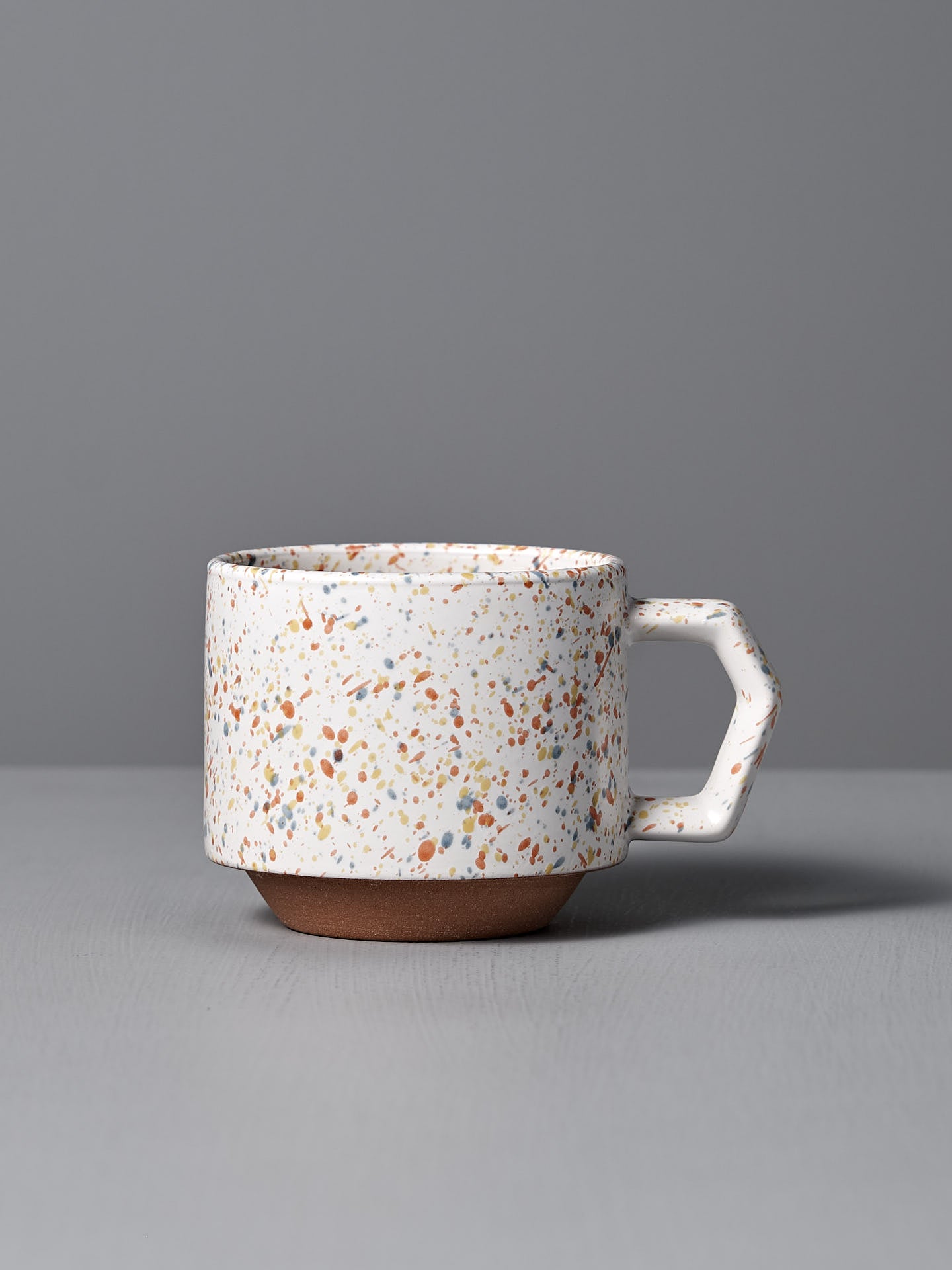 The Stacking Mug – Speckled White, a beautifully crafted piece by CHIPS Inc. artisans, boasts an elegant geometric handle. It is made from speckled Minoyaki porcelain and sits gracefully on a gray surface against a matching gray background.