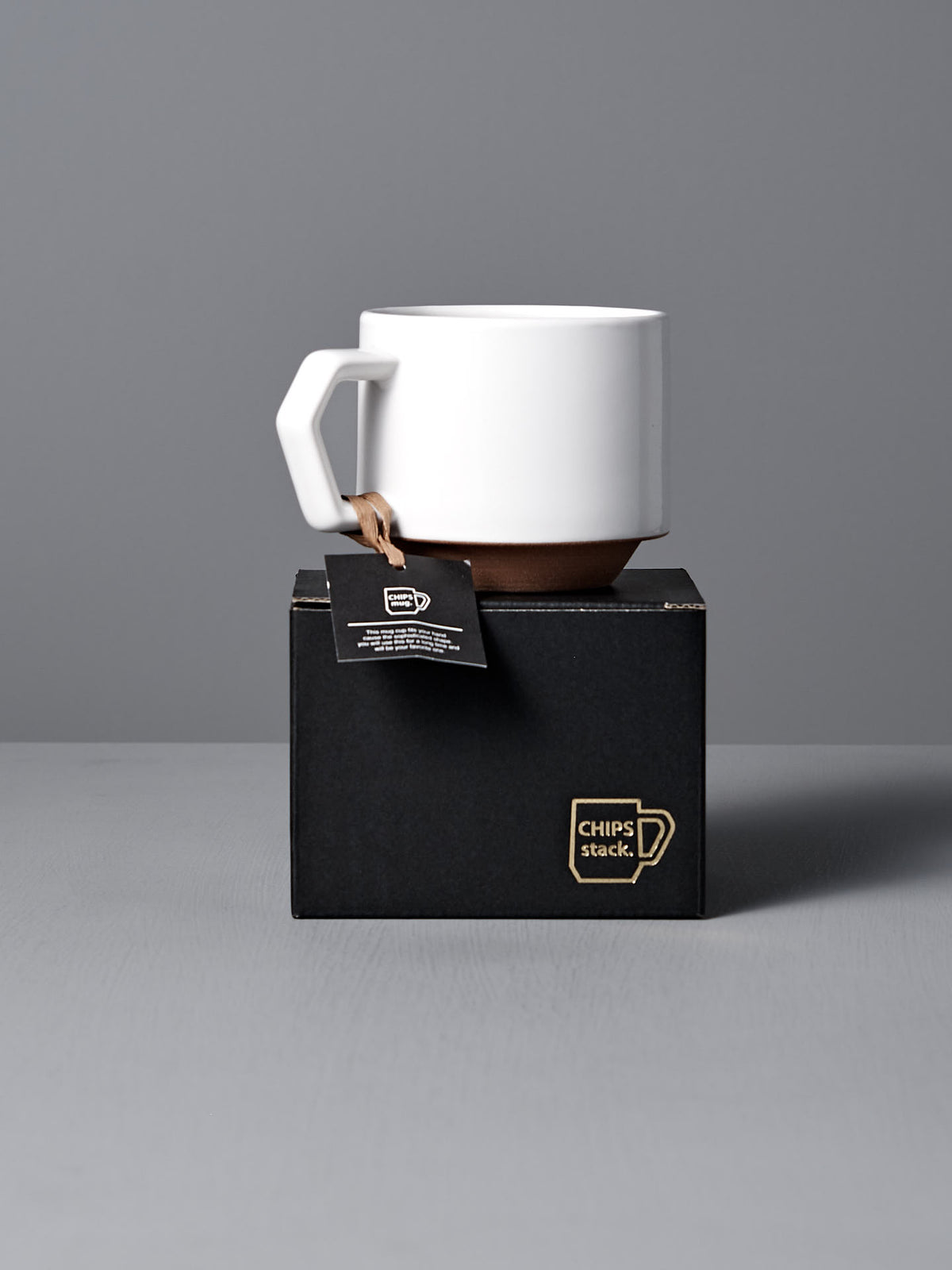 A Stacking Mug – White by CHIPS Inc. with an angular handle sits on a black box labeled &quot;CHIPS stack,&quot; against a neutral background. The design is elegant and stackable, crafted in the CHIPS Minoyaki style, with a tag attached to the mug&#39;s handle.