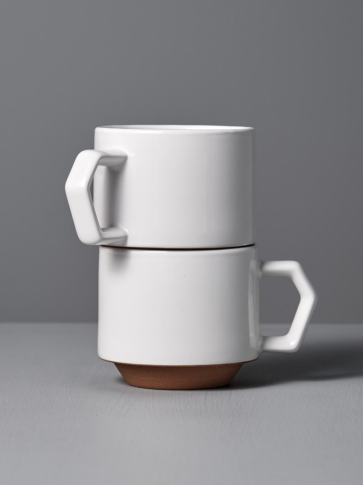 Two Stacking Mug – White from CHIPS Inc., featuring an elegant ceramic design with angular handles, are neatly arranged on a grey surface against a serene grey background.