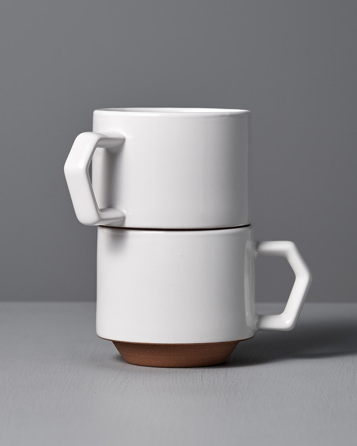 Two Stacking Mug – White from CHIPS Inc., featuring an elegant ceramic design with angular handles, are neatly arranged on a grey surface against a serene grey background.