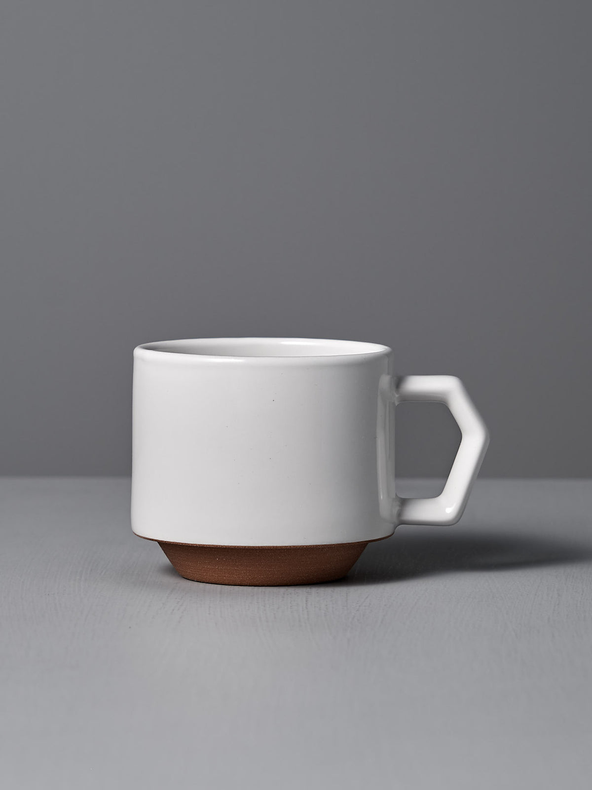 A CHIPS Inc. Stacking Mug – White, featuring a geometric handle and a brown base, sits elegantly on a gray surface against a gray background.