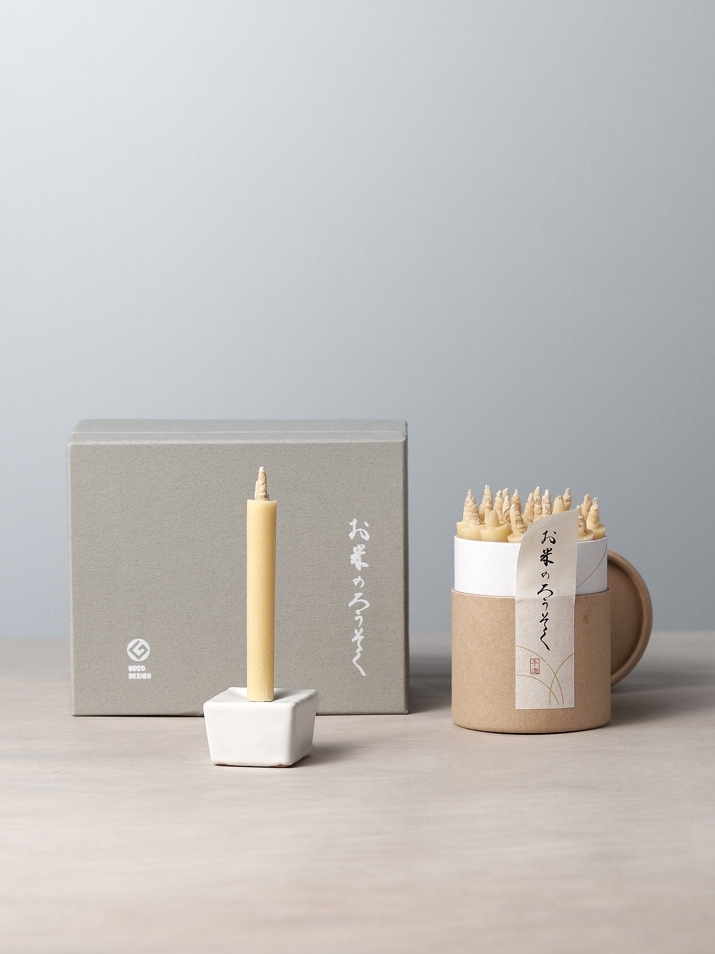 A decorative rice wax candle from the Daiyo brand is elegantly perched in a small holder, accompanied by a cardboard container containing similar candles. In the background, a gray gift box set featuring Japanese writing completes the refined display. This is part of the Rice Wax Candle & Iron Stand 20pc Box Set – Black ⋄ White ⋄ Beige.