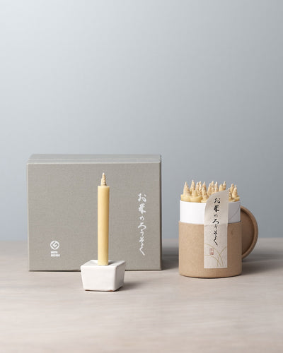 A decorative rice wax candle from the Daiyo brand is elegantly perched in a small holder, accompanied by a cardboard container containing similar candles. In the background, a gray gift box set featuring Japanese writing completes the refined display. This is part of the Rice Wax Candle & Iron Stand 20pc Box Set – Black ⋄ White ⋄ Beige.