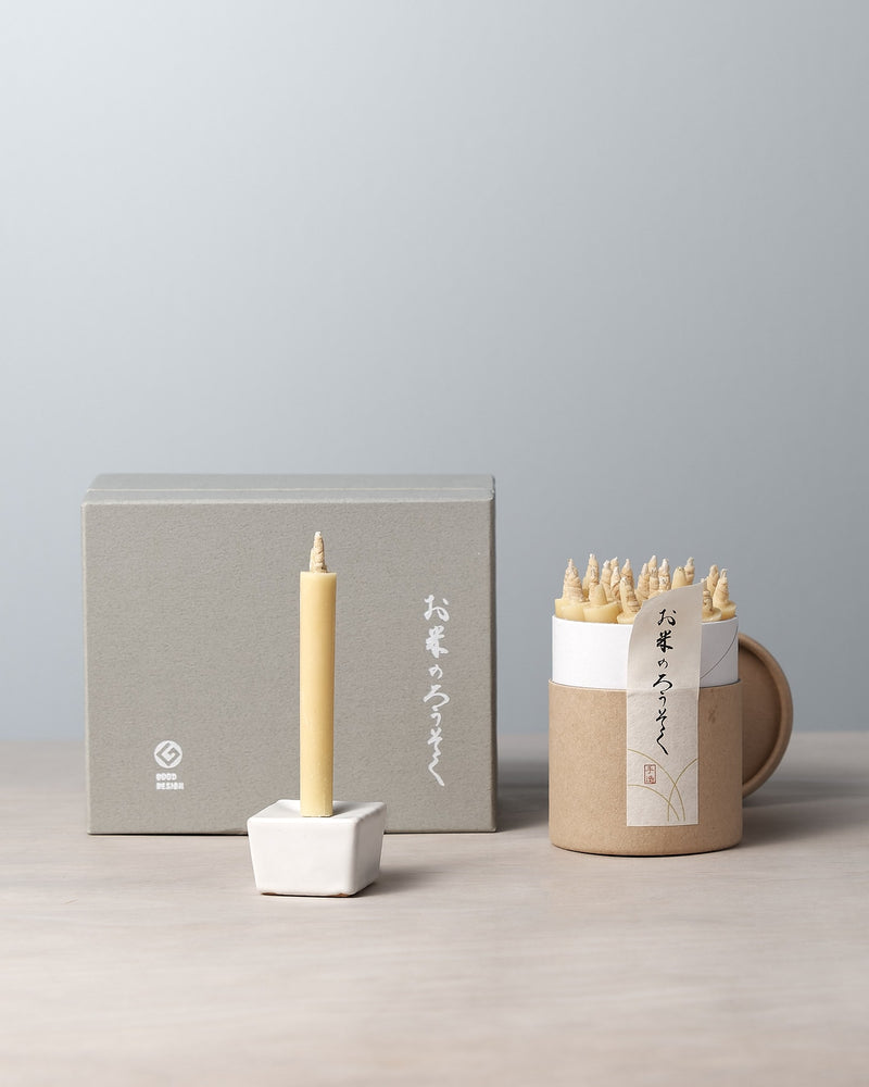 A decorative rice wax candle from the Daiyo brand is elegantly perched in a small holder, accompanied by a cardboard container containing similar candles. In the background, a gray gift box set featuring Japanese writing completes the refined display. This is part of the Rice Wax Candle & Iron Stand 20pc Box Set – Black ⋄ White ⋄ Beige.