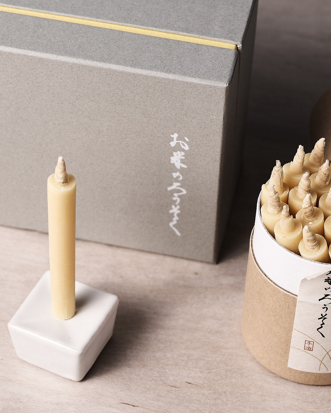 A decorative rice wax candle from the Daiyo brand is elegantly perched in a small holder, accompanied by a cardboard container containing similar candles. In the background, a gray gift box set featuring Japanese writing completes the refined display. This is part of the Rice Wax Candle & Iron Stand 20pc Box Set – Black ⋄ White ⋄ Beige.