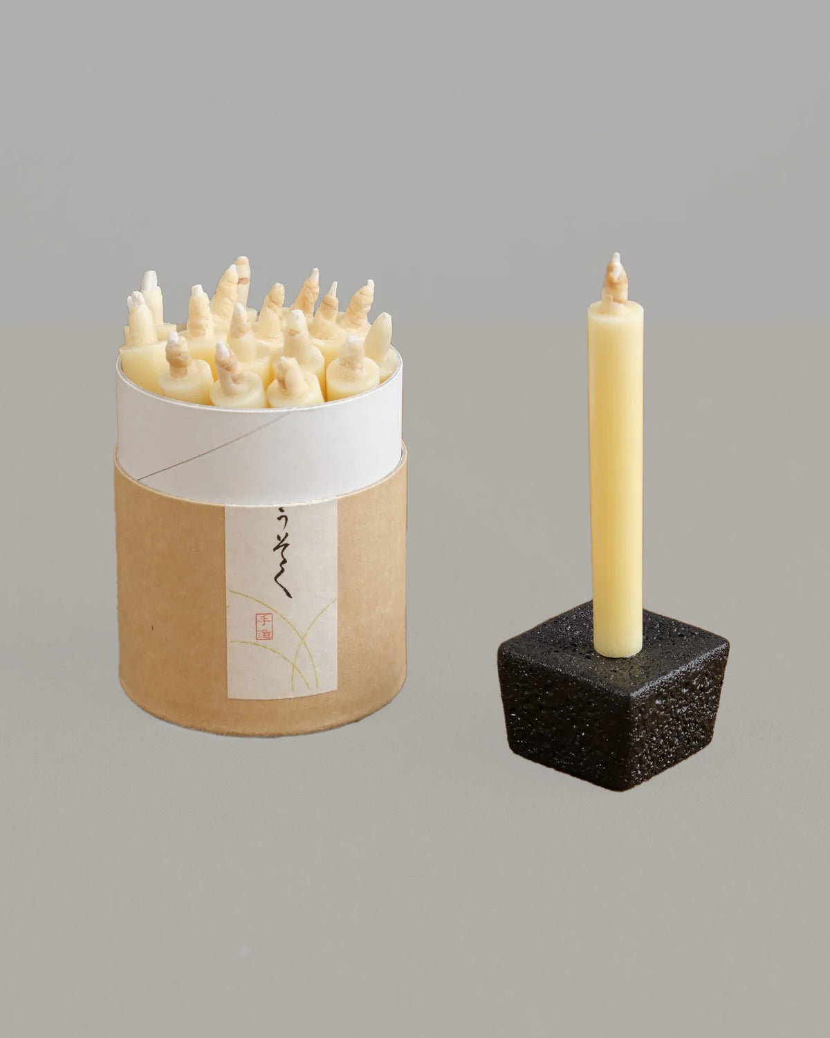 A beige cylindrical container from Daiyo’s Rice Wax Candle & Iron Stand 20pc Box Set in Black, White, and Beige contains spiral rice wax candles, along with a single tall candle displayed in a black Rippoh iron stand, all placed against a light gray background.