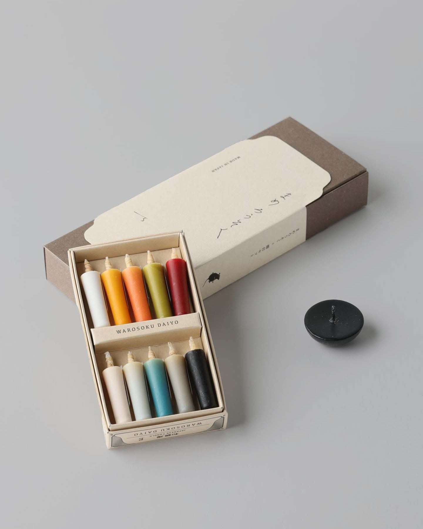 A box containing eight vibrant rice wax candles by Daiyo comes with a white label, each candle in an array of colors. Alongside the box, the Rice Wax Candle & Ceramic Stand Set – Earth | Black sits gracefully on the gray surface, inviting a moment of peace.