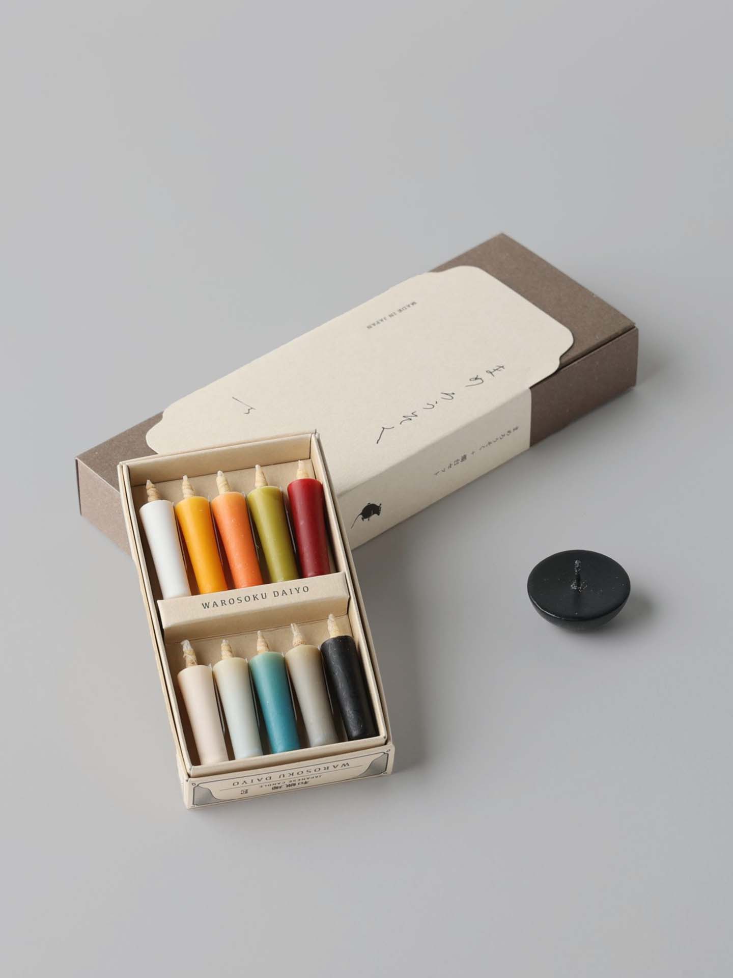 A box containing eight vibrant rice wax candles by Daiyo comes with a white label, each candle in an array of colors. Alongside the box, the Rice Wax Candle & Ceramic Stand Set – Earth | Black sits gracefully on the gray surface, inviting a moment of peace.