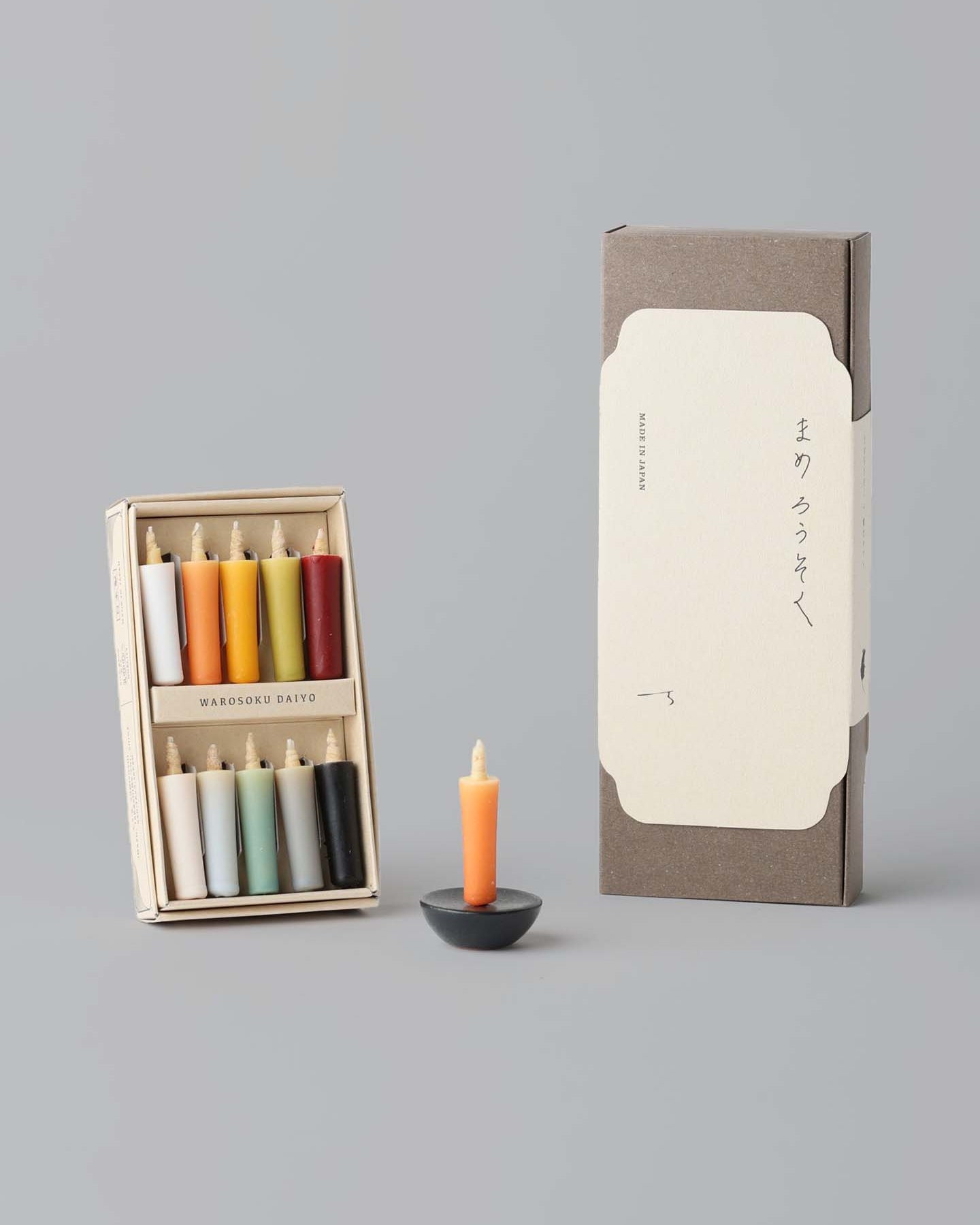 A box containing eight vibrant rice wax candles by Daiyo comes with a white label, each candle in an array of colors. Alongside the box, the Rice Wax Candle & Ceramic Stand Set – Earth | Black sits gracefully on the gray surface, inviting a moment of peace.
