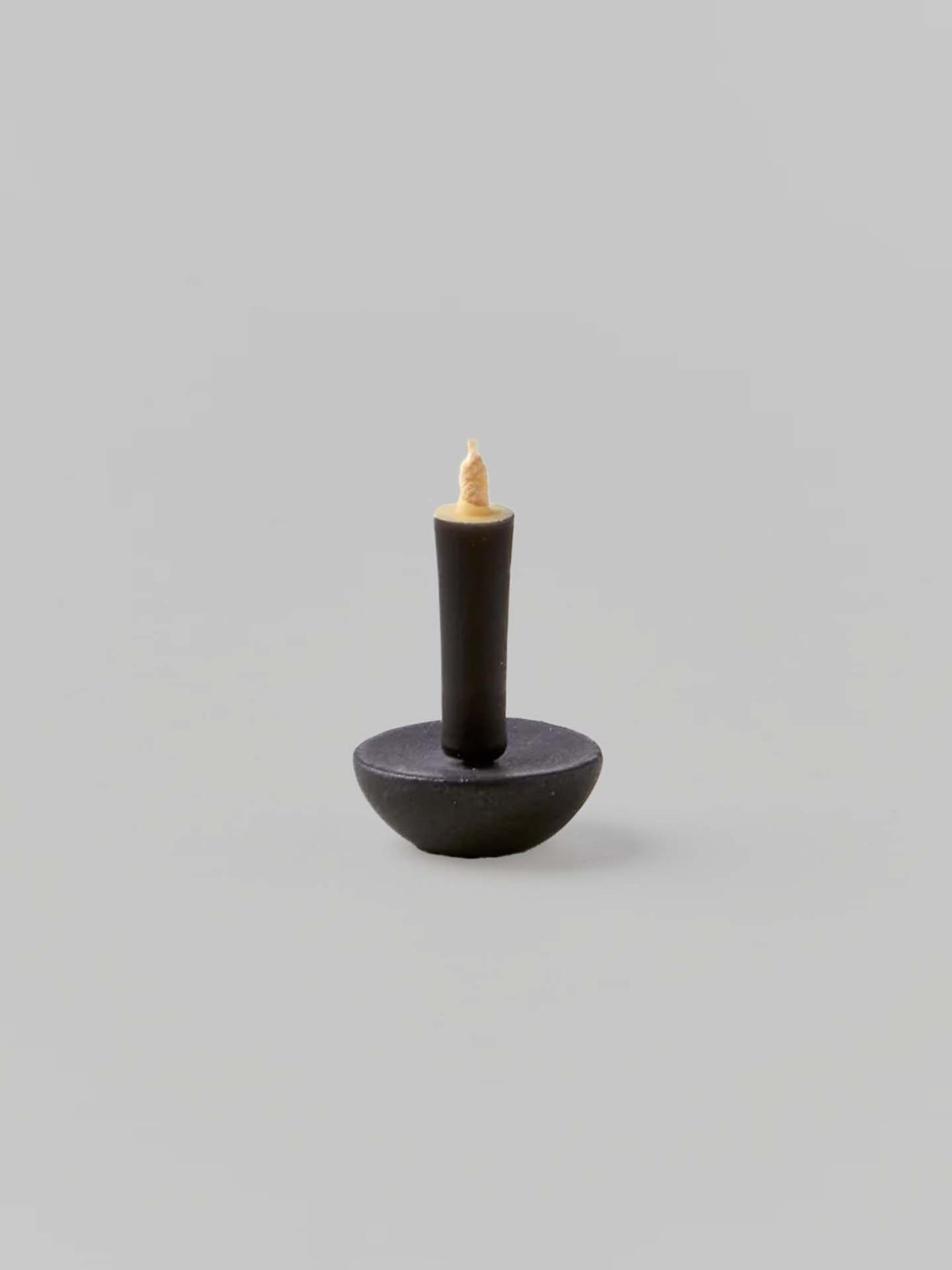 A black candle with a round base and a short, beige wick is elegantly displayed on a gray background. Made by Takazawa Candles, it embodies simplicity and pairs perfectly with the Ceramic Candle Holder Circle – Black from Daiyo, inspired by Shiga prefecture&#39;s ceramic craftsmanship.