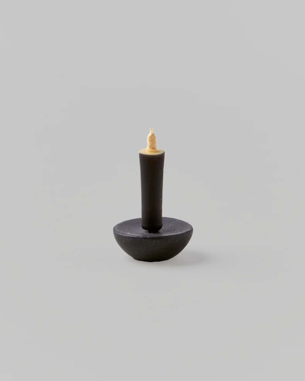 A black candle with a round base and a short, beige wick is elegantly displayed on a gray background. Made by Takazawa Candles, it embodies simplicity and pairs perfectly with the Ceramic Candle Holder Circle – Black from Daiyo, inspired by Shiga prefecture's ceramic craftsmanship.