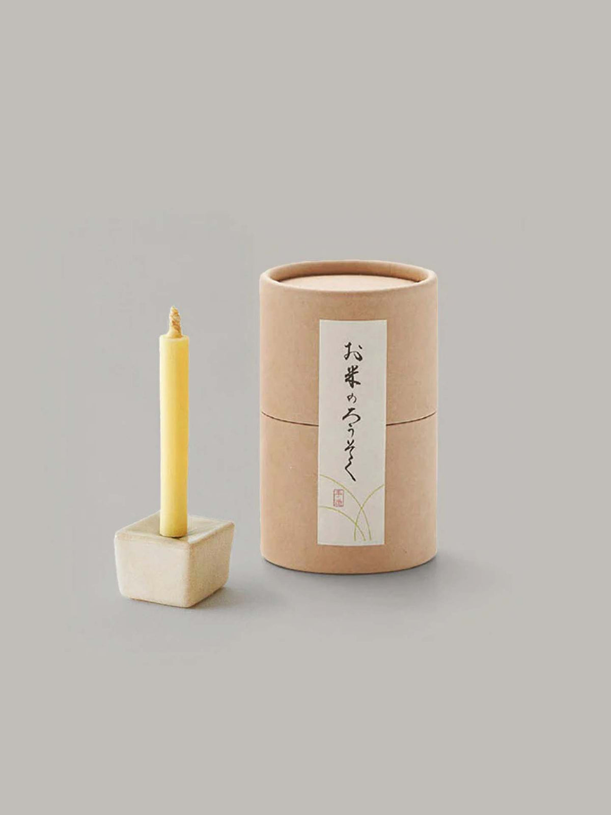 A tall and slender candle made from rice wax is beautifully displayed on an elegant iron and ceramic stand, accompanied by a cylindrical cardboard box adorned with Japanese writing. This stylish arrangement is part of the sophisticated Rice Wax Candle &amp; Iron Stand 20pc Box Set – Black ⋄ White ⋄ Beige by Daiyo, making it perfect for any occasion.