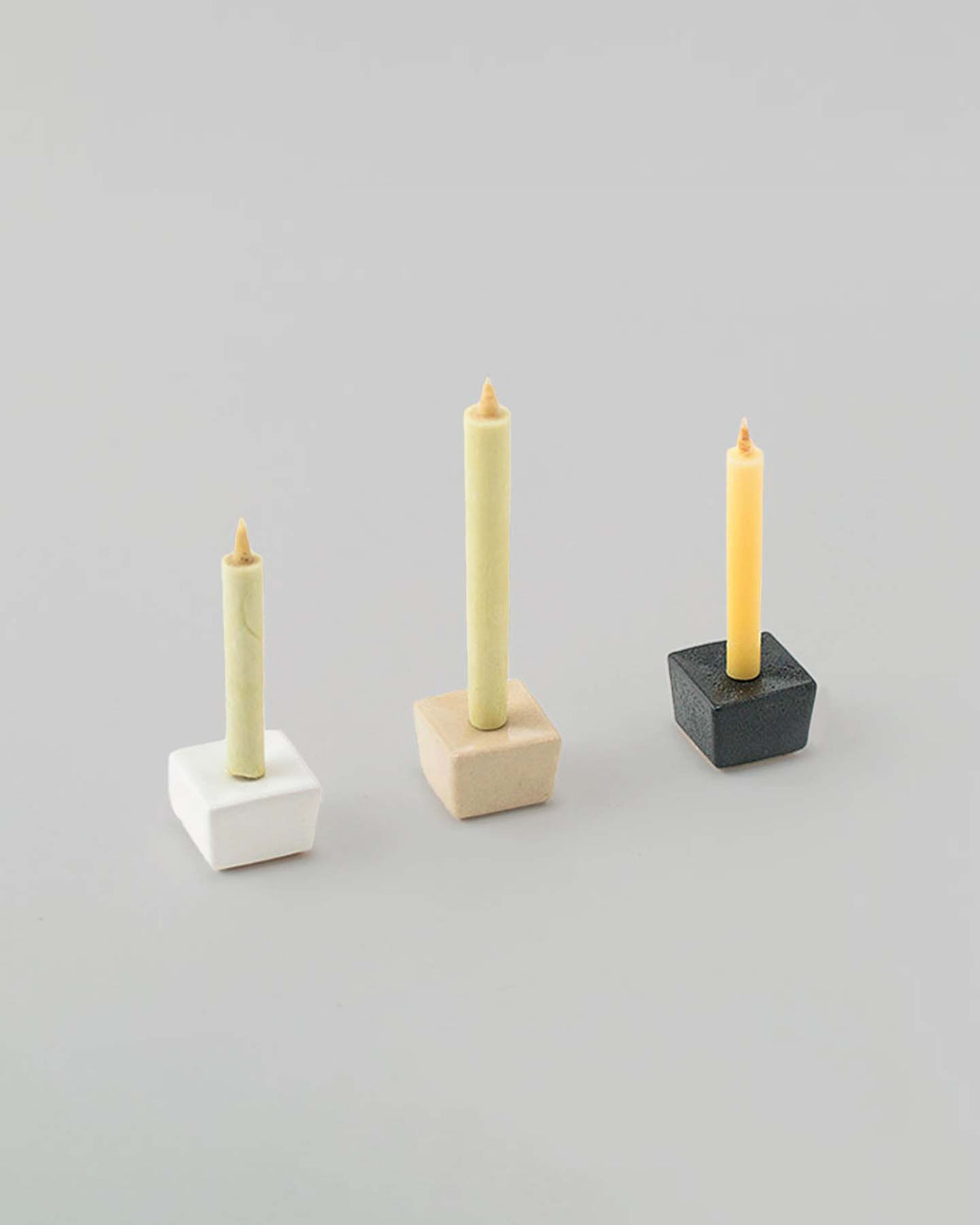 A sophisticated display featuring three Daiyo rice wax candles in modern geometric holders—one in white, one in beige, and one in black—set against a simple light gray background. This elegant arrangement serves as both a stunning presentation and a delightful Rice Wax Candle & Iron Stand 20pc Box Set gift from Daiyo.