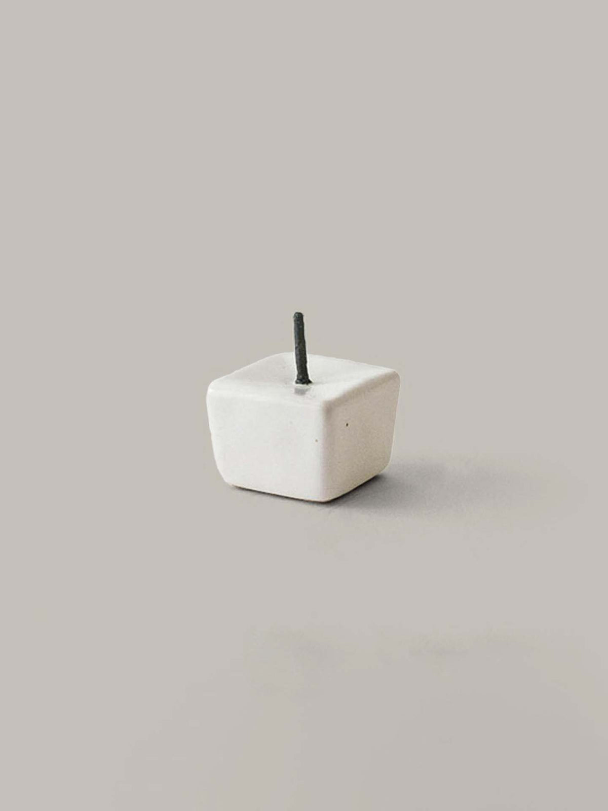 Against a neutral background, there sits a small, cube-shaped item resembling a gift box from the Daiyo Rice Wax Candle &amp; Iron Stand 20pc Box Set – Black ⋄ White ⋄ Beige, featuring a short black stick emerging from its top.