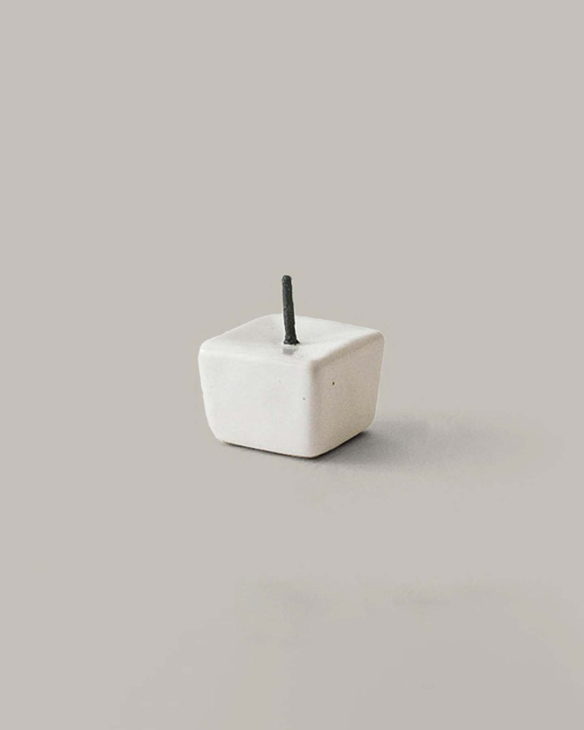 Against a neutral background, there sits a small, cube-shaped item resembling a gift box from the Daiyo Rice Wax Candle & Iron Stand 20pc Box Set – Black ⋄ White ⋄ Beige, featuring a short black stick emerging from its top.