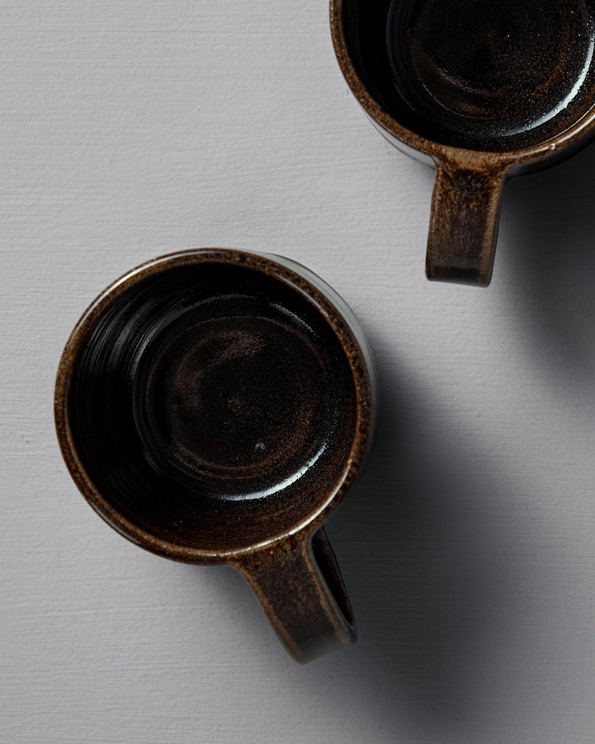 Two empty Studio Foon Everyday Stacker Cups – Medium in Burnt Umber, handmade and dark brown, sit on a light gray textured surface—ideal for elevating your daily coffee ritual.