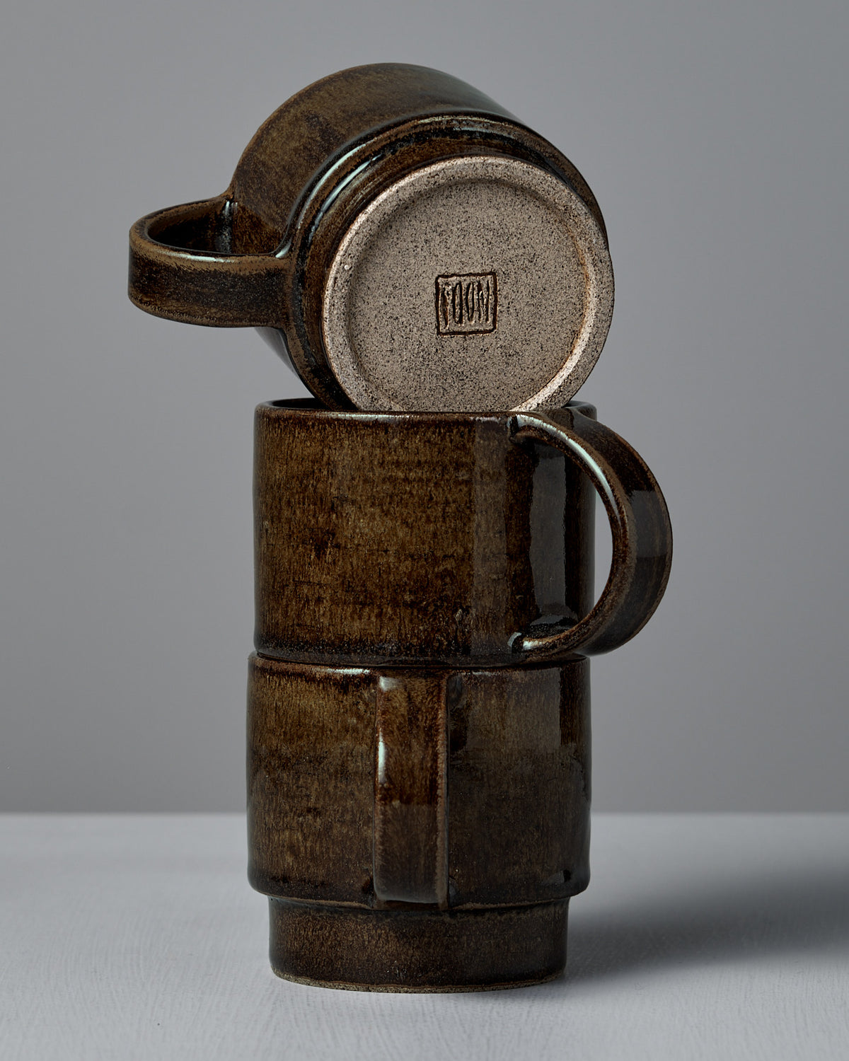 Two stacked Studio Foon Everyday Stacker Cups – Large in Burnt Umber, with the top cup upside down to display the maker's mark, set against a plain gray background.