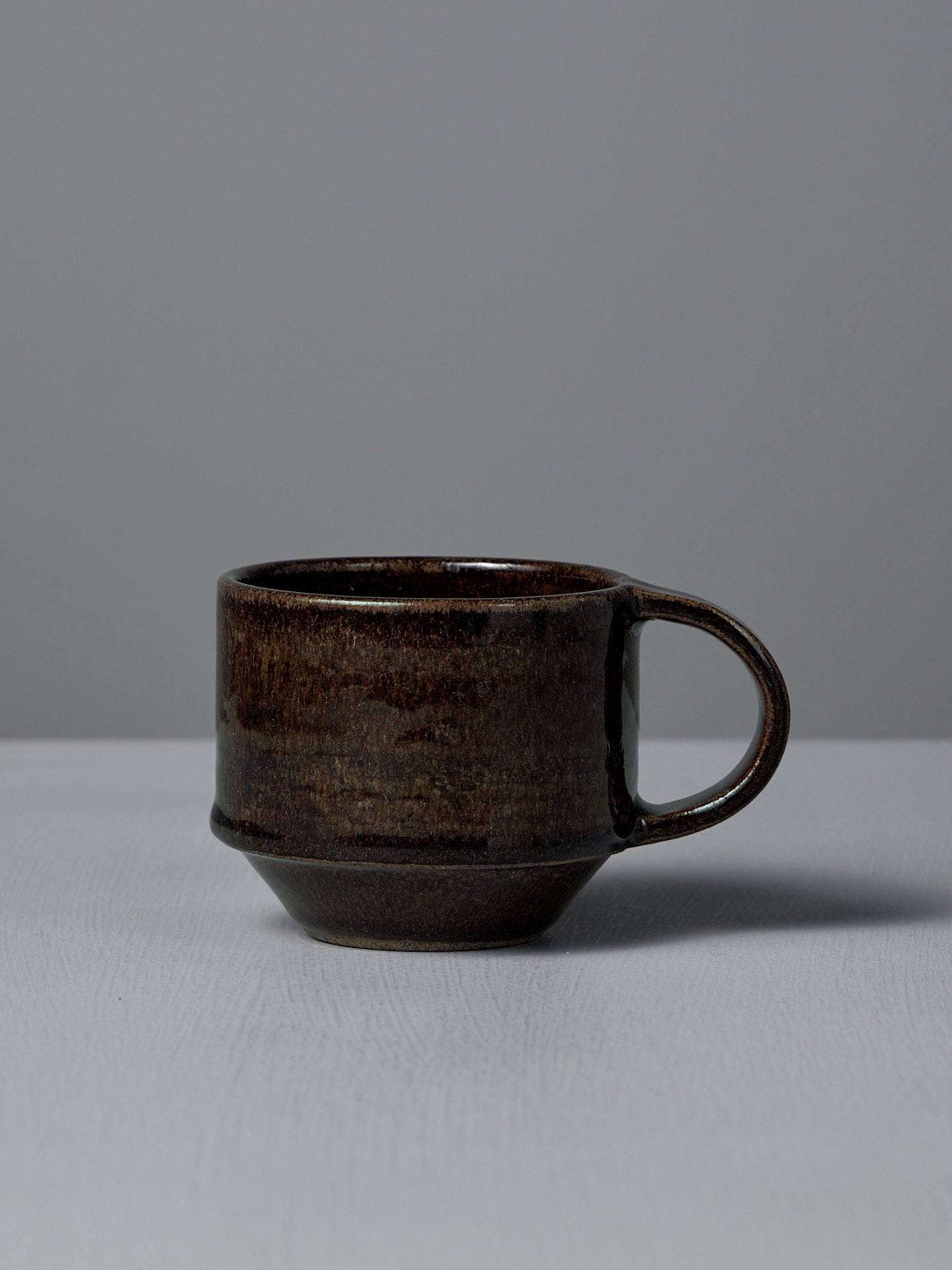 The Studio Foon Everyday Stacker Cup – Medium in Burnt Umber, a dark brown handcrafted cup with a large handle, rests on a light gray surface against a plain gray background.