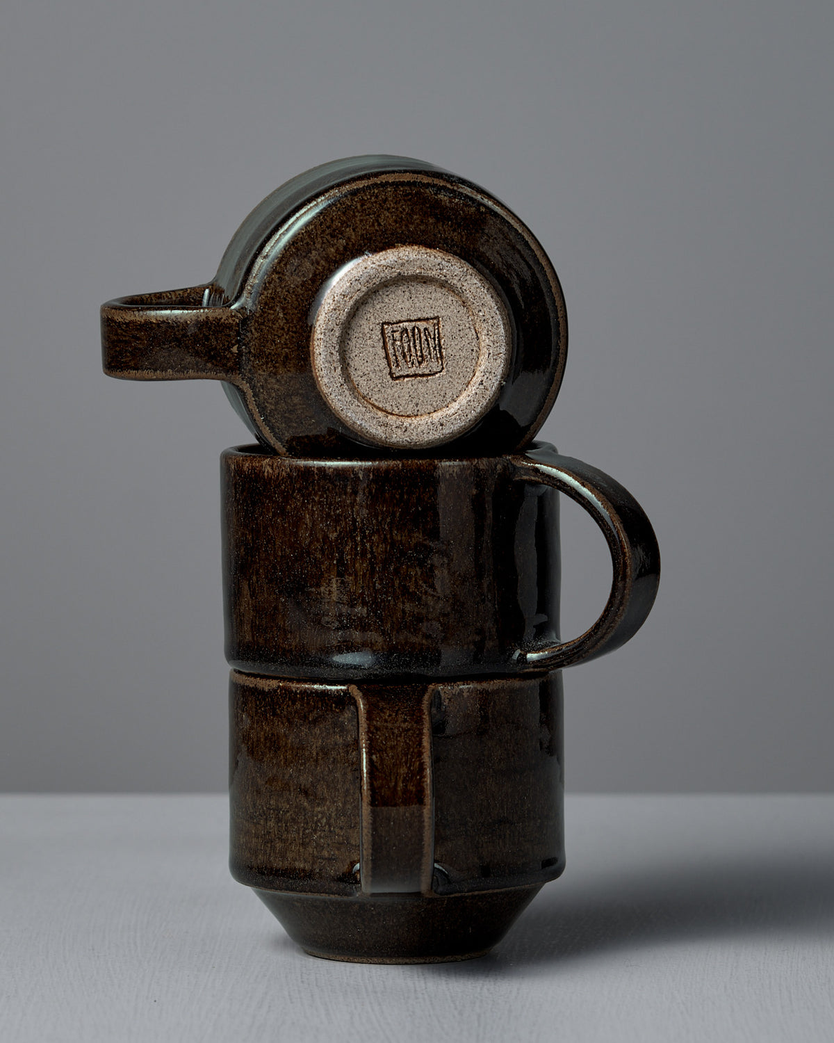 Two Studio Foon Everyday Stacker Cups in Burnt Umber are stacked vertically, with the top medium cup upside down to display its stamped base logo.
