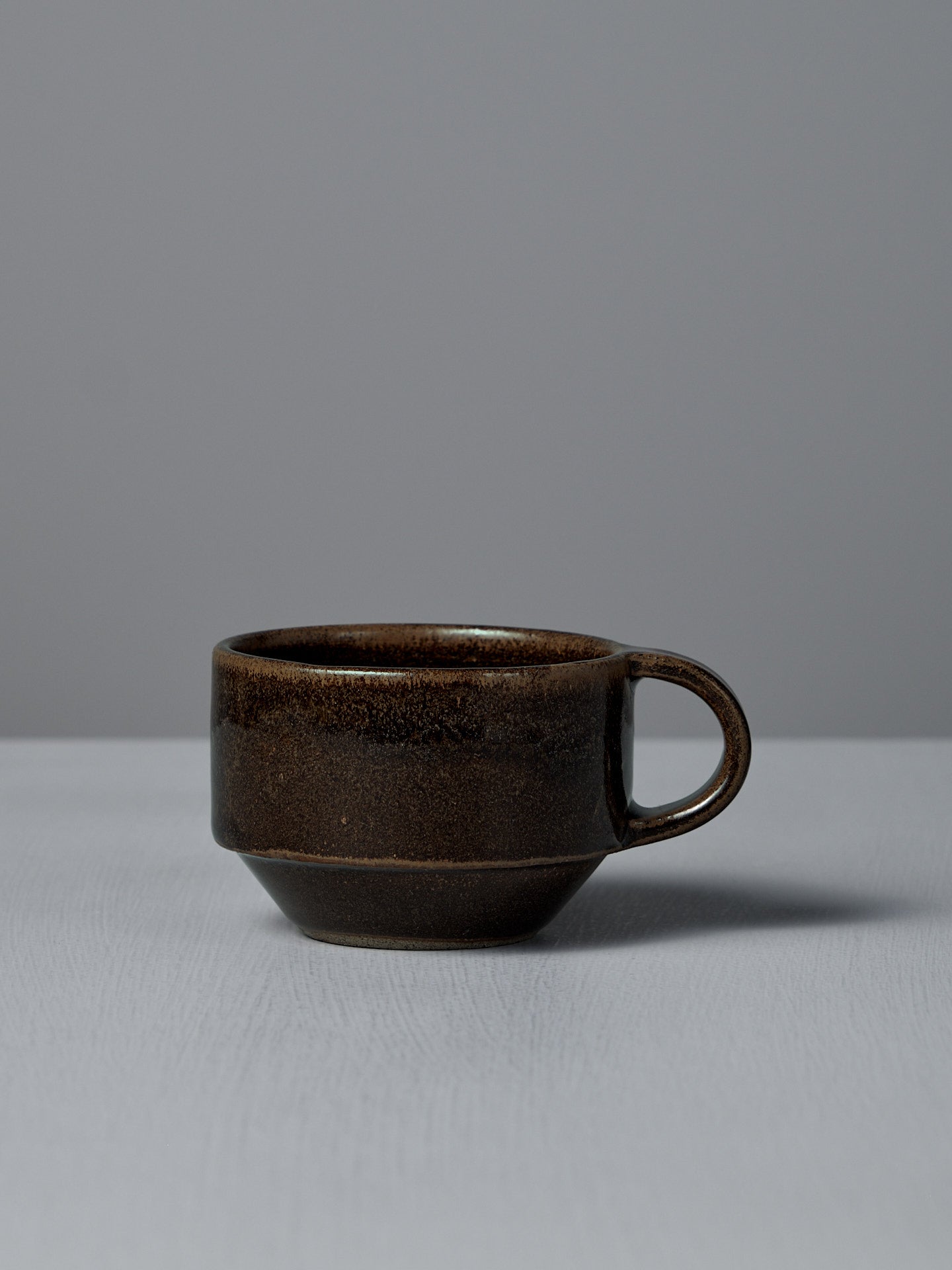 The Studio Foon Everyday Stacker Cup – Small in Burnt Umber, a dark brown ceramic cup with handle, rests on a light gray surface against a plain gray background, showcasing its charming minimalist design.