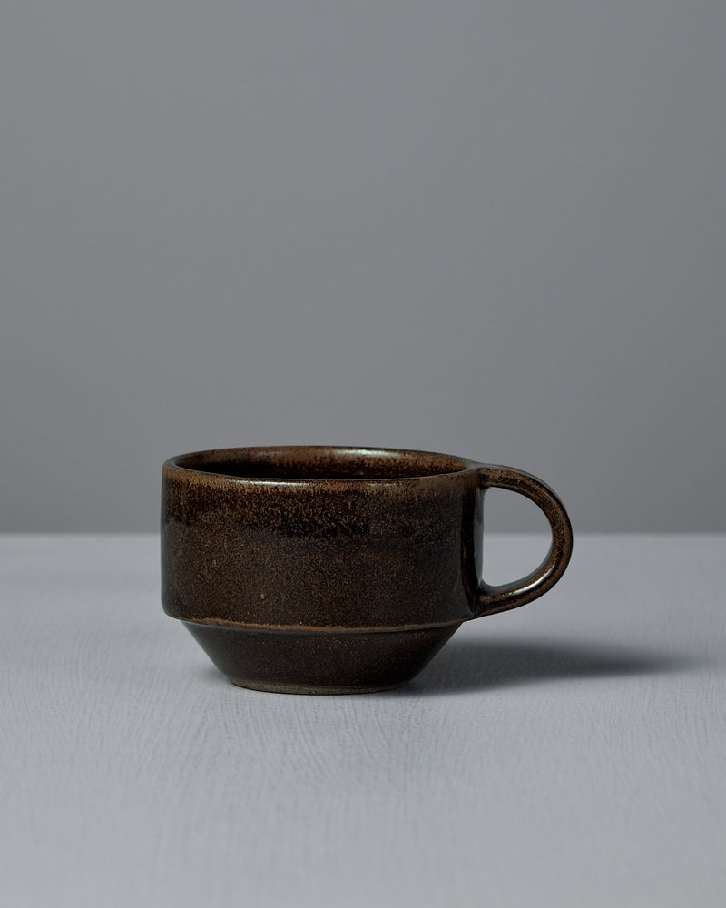 The Studio Foon Everyday Stacker Cup – Small in Burnt Umber, a dark brown ceramic cup with handle, rests on a light gray surface against a plain gray background, showcasing its charming minimalist design.
