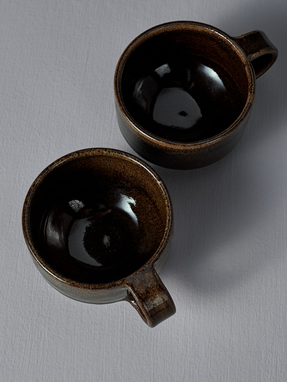 Two empty Studio Foon Everyday Stacker Cups – Small in Burnt Umber, handcrafted with angled handles, rest on a light grey textured surface.