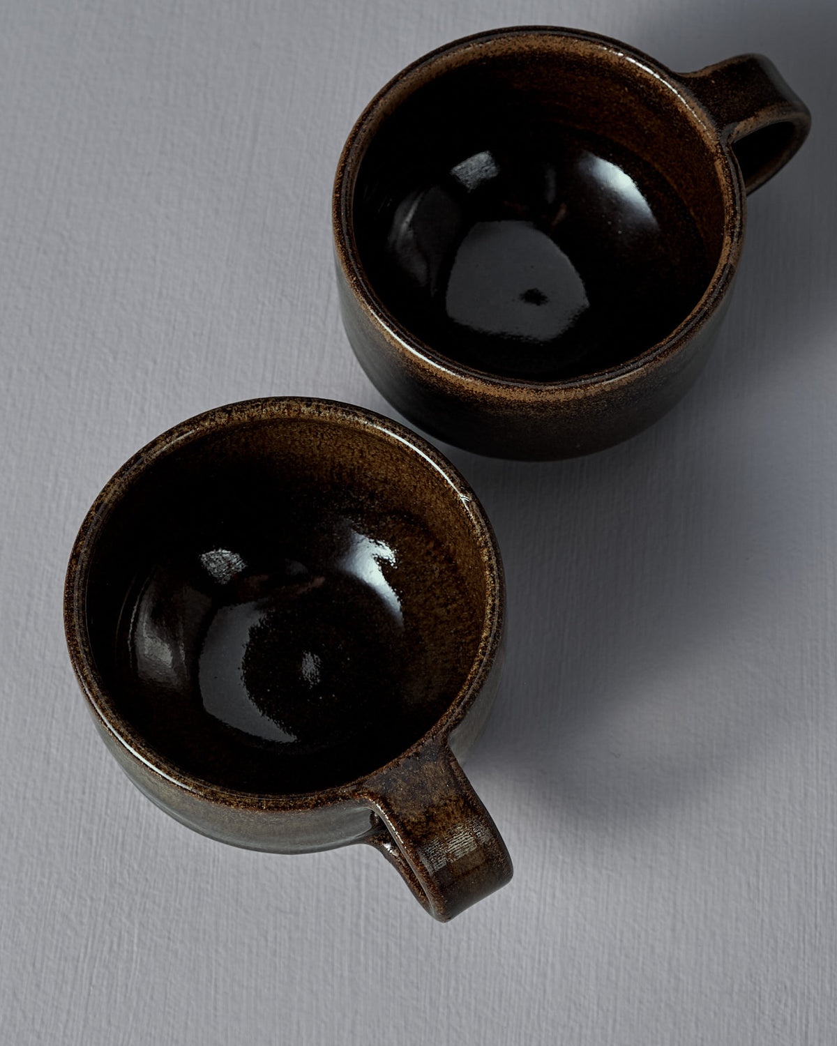 Two empty Studio Foon Everyday Stacker Cups – Small in Burnt Umber, handcrafted with angled handles, rest on a light grey textured surface.