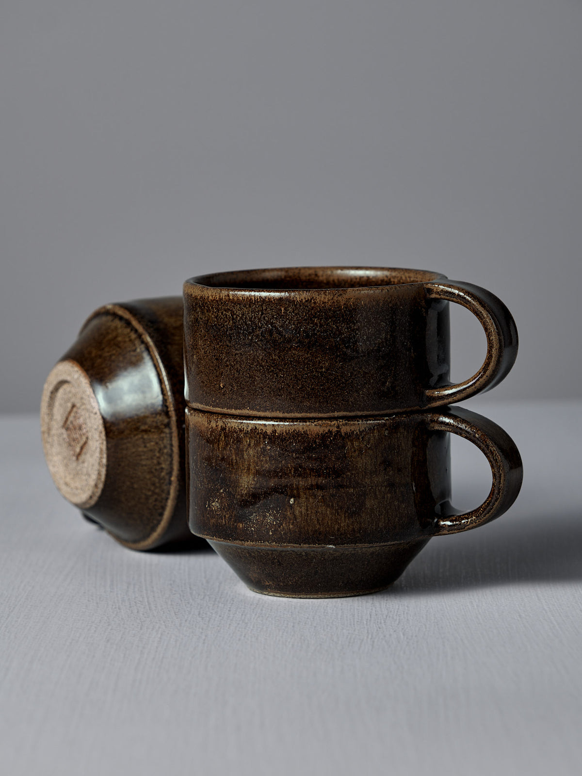 Two Studio Foon Everyday Stacker Cups in Burnt Umber are stacked, with a third cup lying on its side in the background. All are set against a plain grey surface and wall.